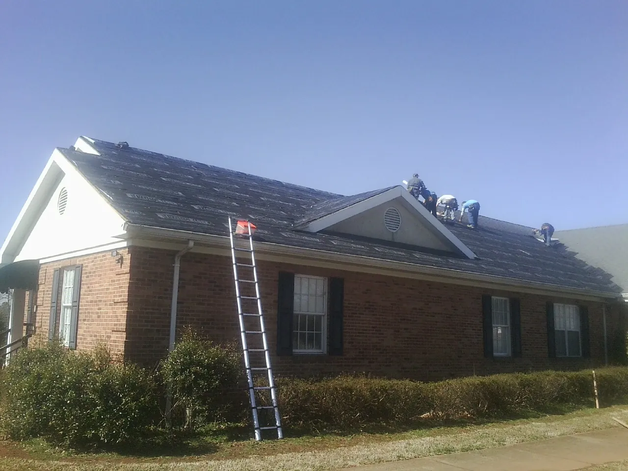 Crew tearing shingles off a brick ranch roof with a ladder set — Metal Roof Repair work by our crew in Bohemia