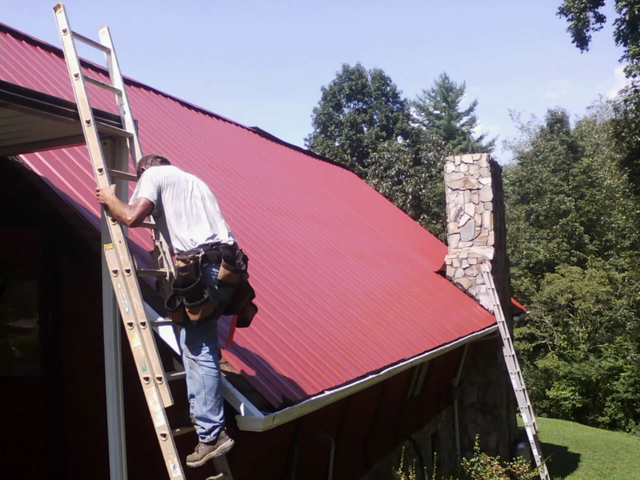 Roofer on a ladder beside a red metal roof and stone chimney — Soffit & Fascia Repair by Roofing Brothers in Bohemia